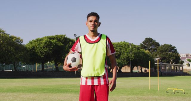 Young male soccer player standing on field, holding soccer ball with confident expression, wearing green training vest and red shorts. Ideal for use in sports-related articles, training tutorials, advertisements for sportswear or upcoming events, and motivational content.