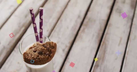 Glass of milk placed on weathered wooden table with chocolate chip cookie resting on top, through two purple-striped paper straws sticking out. Bright, pastel-colored geometric shapes sprinkling around to add whimsical touch. Useful for advertisements focusing on classic snack themes, dairy promotions, or playful rustic kitchen settings.
