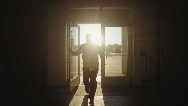 Walking backlit man passing through glass doors into sunlit parking lot with lens flare