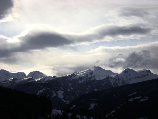 This photo depicts a dramatic sky with thick clouds over a snow-capped mountain range. Ideal for uses in travel or nature magazines, winter-themed advertisements, or as a stunning background in presentations that require a captivating natural setting.