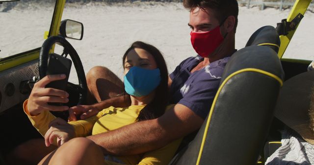 Happy caucasian couple wearing face masks sitting in beach buggy taking selfies. beach stop off on summer holiday road trip during coronavirus covid 19 pandemic.