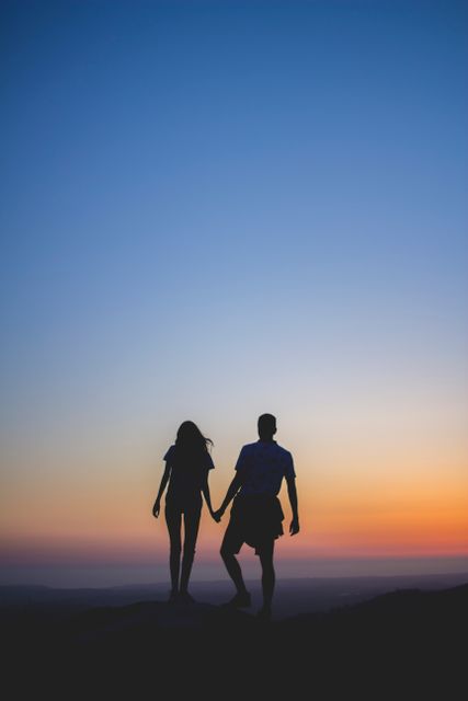 Silhouette of couple holding hands during sunset on hilltop. Ideal for use in romantic, travel, and adventure-themed advertisements, blogs about relationships, or articles highlighting the beauty of nature and connecting with loved ones.