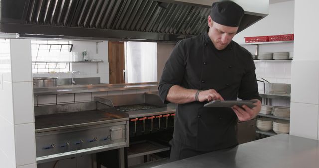 Professional chef standing in a commercial kitchen using a digital tablet. Ideal for concepts related to modern kitchen technology, restaurant management, cooking, culinary arts, professional chefs, and efficient kitchen operations. Can be used in articles, blog posts, and advertisements for kitchen equipment, restaurant technology, and culinary training programs.