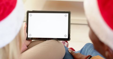 Diverse couple wearing santa hats holding tablet with blank screen while relaxing on couch