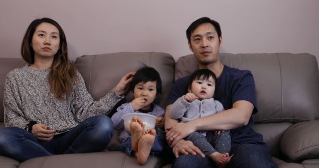 Asian family with two young children sitting on a couch in a living room, displaying relaxed home life. Parents look attentive, with mother stroking her child’s hair. This image can be used for themes around family, togetherness, home comfort, and parenting.