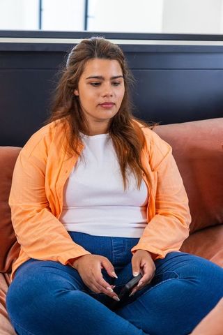 Adult african woman sitting on sofa holding smartphone wearing orange shirt and jeans