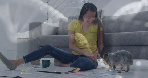 Smiling korean woman sitting on living room floor petting small dog with laptop