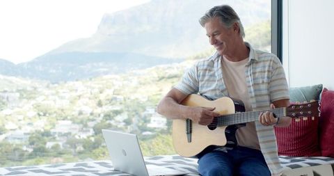 Smiling mature man playing acoustic guitar on window bench overlooking mountain town