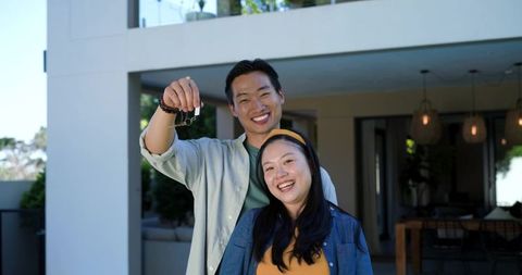 Young asian couple celebrating new home while holding keys outside modern house balcony
