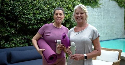 Mature women standing by backyard pool holding purple yoga mats and water bottles