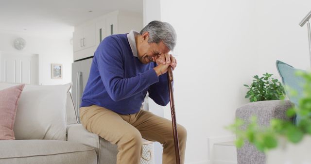 Elderly man in blue sweater and beige pants resting on a cane, sitting thoughtfully in a bright living room. Suitable for use in content related to aging, senior living, emotions, solitude, and life challenges. Can be used in articles about elderly care, mental health, introspection, and personal stories of older adults.