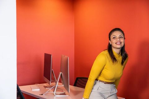 Confident woman smiling and leaning on white desk with silver monitors against bright orange wall