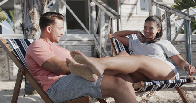 Young couple sitting comfortably on deck chairs on a sunny beach. They are engaged in conversation, smiling, and enjoying each other's company. Ideal for use in travel promotions, vacation advertisements, relaxation themes, and lifestyle blog posts about enjoying summer or tropical getaways.