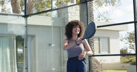Smiling african american woman playing padel on backyard court holding paddle and laughing