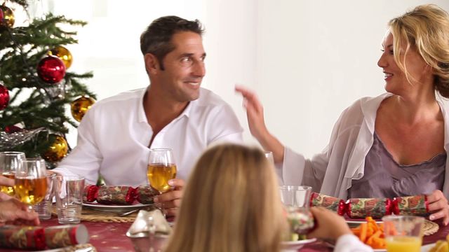 Family enjoying festive Christmas celebration around dinner table. Smiles and conversation create joyful atmosphere near Christmas tree adorned with ornaments. Suitable for holiday greeting cards, family-oriented advertisements, and festive blogs.