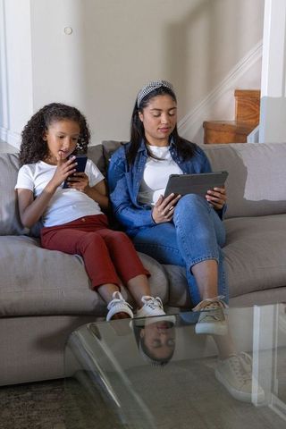 Diverse mother and daughter relaxing on couch, browsing tablet and phone together