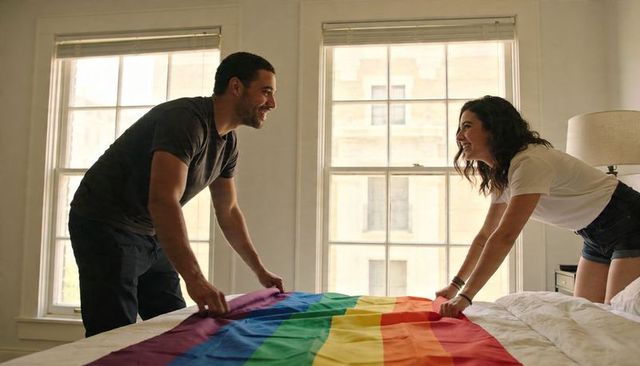 Couple spreading rainbow pride flag on bed, smiling and straightening banner together