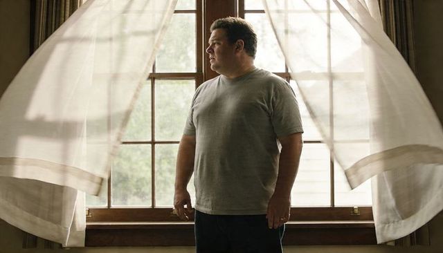 Midlife man standing by window with billowing curtains, wearing wedding band, gazing left, contempla