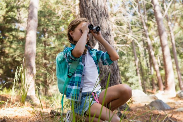 Young girl kneeling in forest, looking through binoculars with a backpack on her back. Ideal for themes of childhood adventure, nature exploration, outdoor activities, and wildlife observation. Perfect for educational materials, travel blogs, and environmental awareness campaigns.