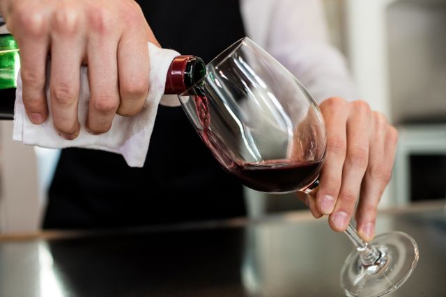 This image shows a barkeeper pouring red wine into a glass at a bar counter. It is ideal for use in articles or advertisements related to hospitality, wine service, restaurants, and professional bartending. It can also be used in content about wine tasting events, sommelier training, and beverage service.