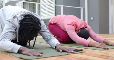 African American friends practicing yoga on sunlit balcony performing child pose together