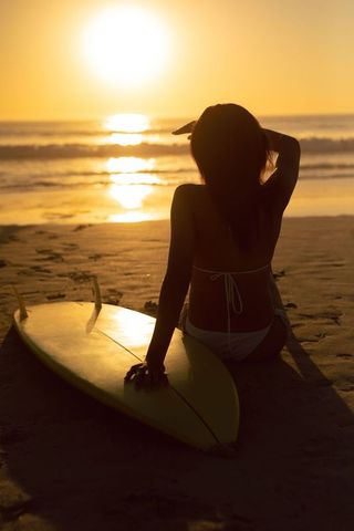African American surfer watching golden sunset sitting on beach with yellow surfboard