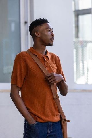 African man wearing rust polo holding brown leather crossbody strap on urban sidewalk