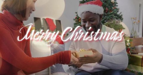 Interracial couple exchanging gold gift on sofa by christmas tree wearing santa hat
