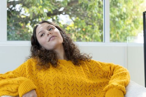 Young woman reclining on sofa in mustard sweater holding clear tumbler sunlit minimal home