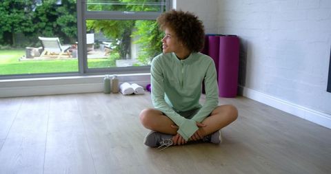 Young african american woman sitting cross-legged gazing out window in sunlit home gym