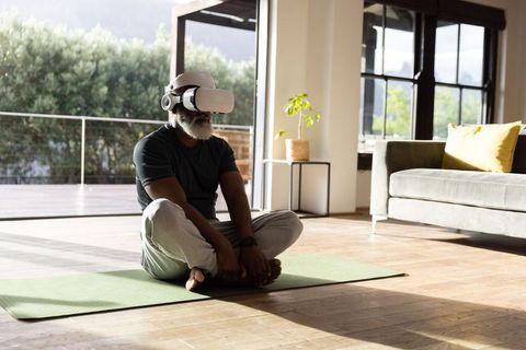 Senior african american man meditating with vr headset on yoga mat in bright living room