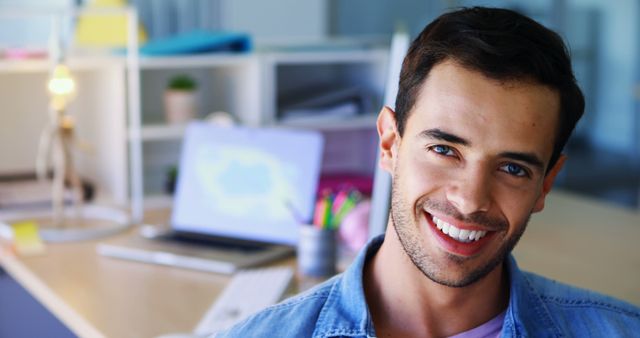 Young professional smiling in front of a computer screen in a modern office setting. Ideal for use in articles about workplace culture, office productivity, and modern work environments. Great for business-related content, career development blogs, and advertising office equipment or workspace solutions.