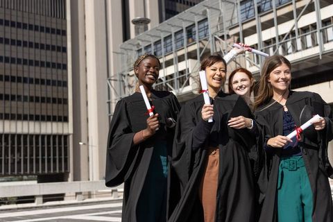 Multicultural female graduates celebrating with diplomas on sunlit plaza by office towers