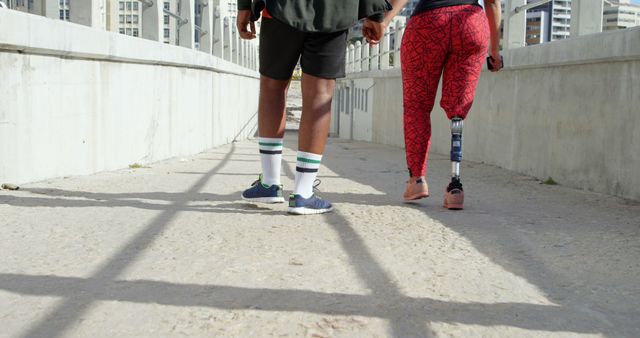 This image depicts a couple walking on an urban pathway, with a focus on their lower bodies. The woman is wearing a prosthetic leg, highlighting themes of diversity, inclusivity, and an active lifestyle. The setting suggests a city environment, making it ideal for use in content related to urban living, fitness, or inspirational and motivational contexts.