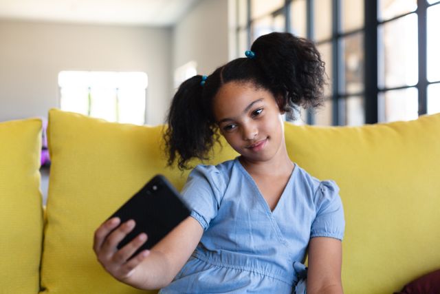 Young biracial girl sitting on a yellow couch, taking a selfie with a smartphone. She is wearing a light blue dress and has her hair in pigtails. This image can be used for educational materials, technology in education, childhood relaxation, and social media content.