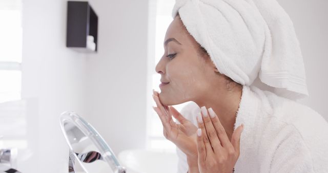 Woman wearing towel on head and bathrobe, applying skincare cream on face, looking in mirror. Ideal for blogs, articles, and advertising related to skincare tips, beauty routines, self-care practices, and healthy skin maintenance.