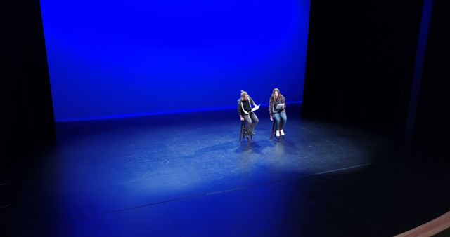 Two young Caucasian females are on a stage under a blue spotlight, with one sitting on a stool and the other standing, rehearsing a performance or dialogue, with copy space. Their positioning and the stark lighting create a dramatic atmosphere, hinting at a theatrical or dance production.