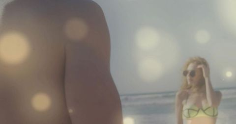 Cropped male torso standing on sunlit beach with woman adjusting sunglasses in neon bikini