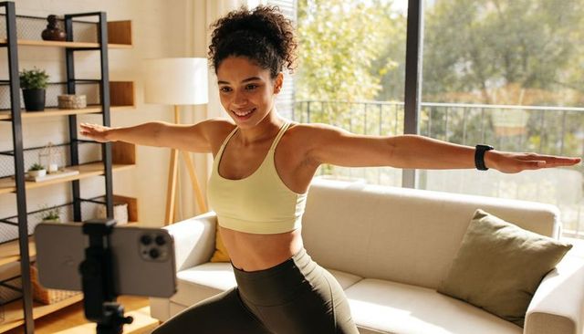 Young woman working out at home with smartphone tripod and smartwatch, yoga lunge tutorial