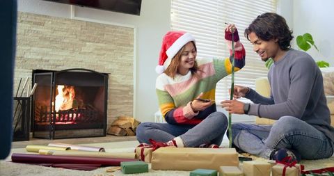 Couple wrapping christmas gifts by fireplace, woman wearing santa hat, cozy holiday scene