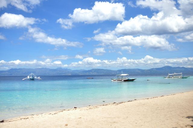 Idyllic tropical beach scene with boats on clear blue water and a sandy shore under a bright sunny sky with fluffy clouds. Perfect for travel brochures, vacation advertisements, and websites promoting tropical holiday destinations.