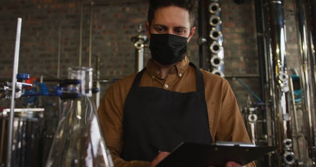 Male worker wearing a black mask and apron, conducting inspection on clipboard in a brewery. Useful for topics related to safety protocols, COVID-19 measures in workplace, industrial inspections, and brewery operations.