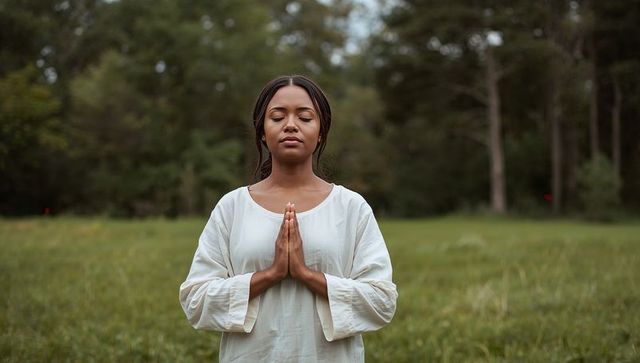 Woman meditating in meadow with hands pressed together wearing white tunic serene outdoor portrait