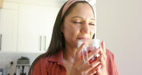 Woman sipping warm tea in bright kitchen holding glass mug close to face