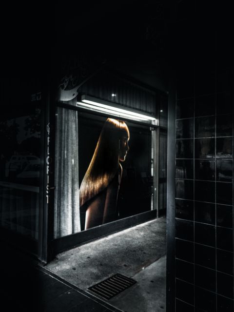 Backlit mannequin through storefront window scene depicts dark, urban setting. Dramatic lighting highlights mannequin's silhouette and glossy appearance. Useful for advertisements regarding nighttime fashion, store window displays, or urban photography art collections.