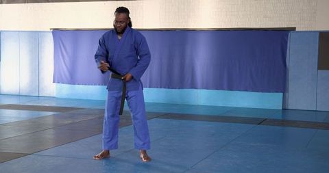 Martial artist in blue gi tying black belt in training hall