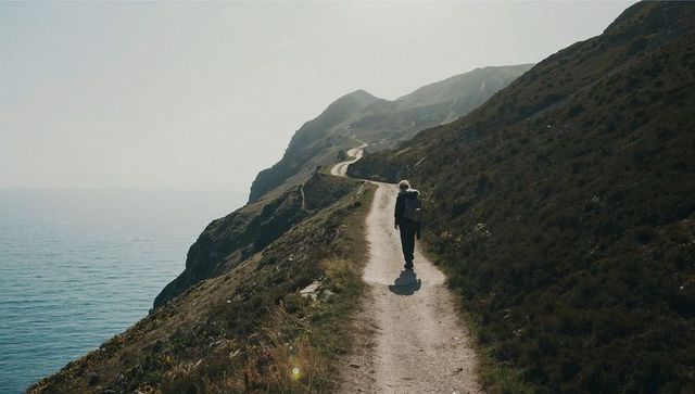 Solitary hiker walking rugged cliffside coastal trail with backpack toward misty horizon
