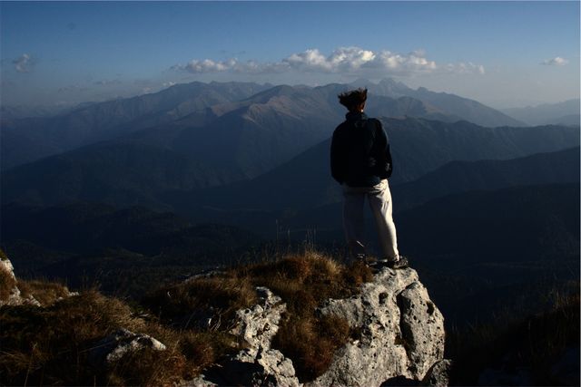 An individual is standing on a rocky ledge, gazing over a vast mountain range with peaks and valleys in the distance. This scene evokes a sense of adventure, freedom, and connection with nature. Useful for content related to outdoor activities, travel inspiration, adventure experiences, and environmental appreciation.