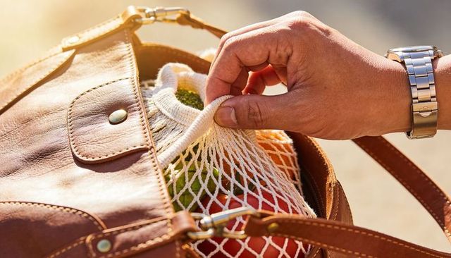 Hand reaching for mesh produce bag inside brown leather handbag in sunlight