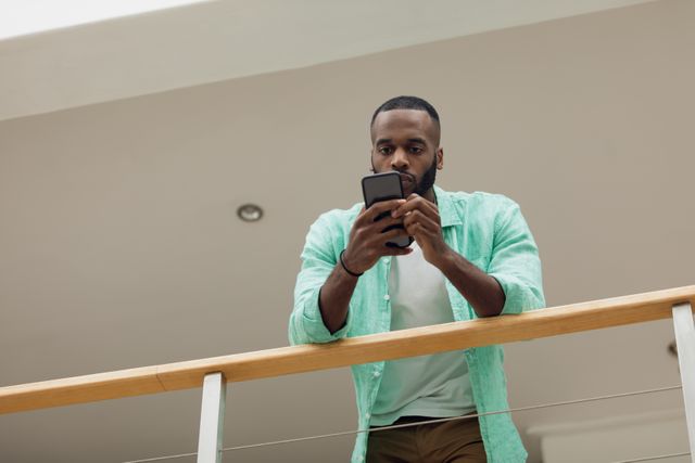 This image depicts an African-American man using a smartphone while leaning on a wooden rail. The low angle view emphasizes his focus and engagement with the device. Ideal for use in articles or advertisements related to technology, communication, modern lifestyle, or mobile applications.